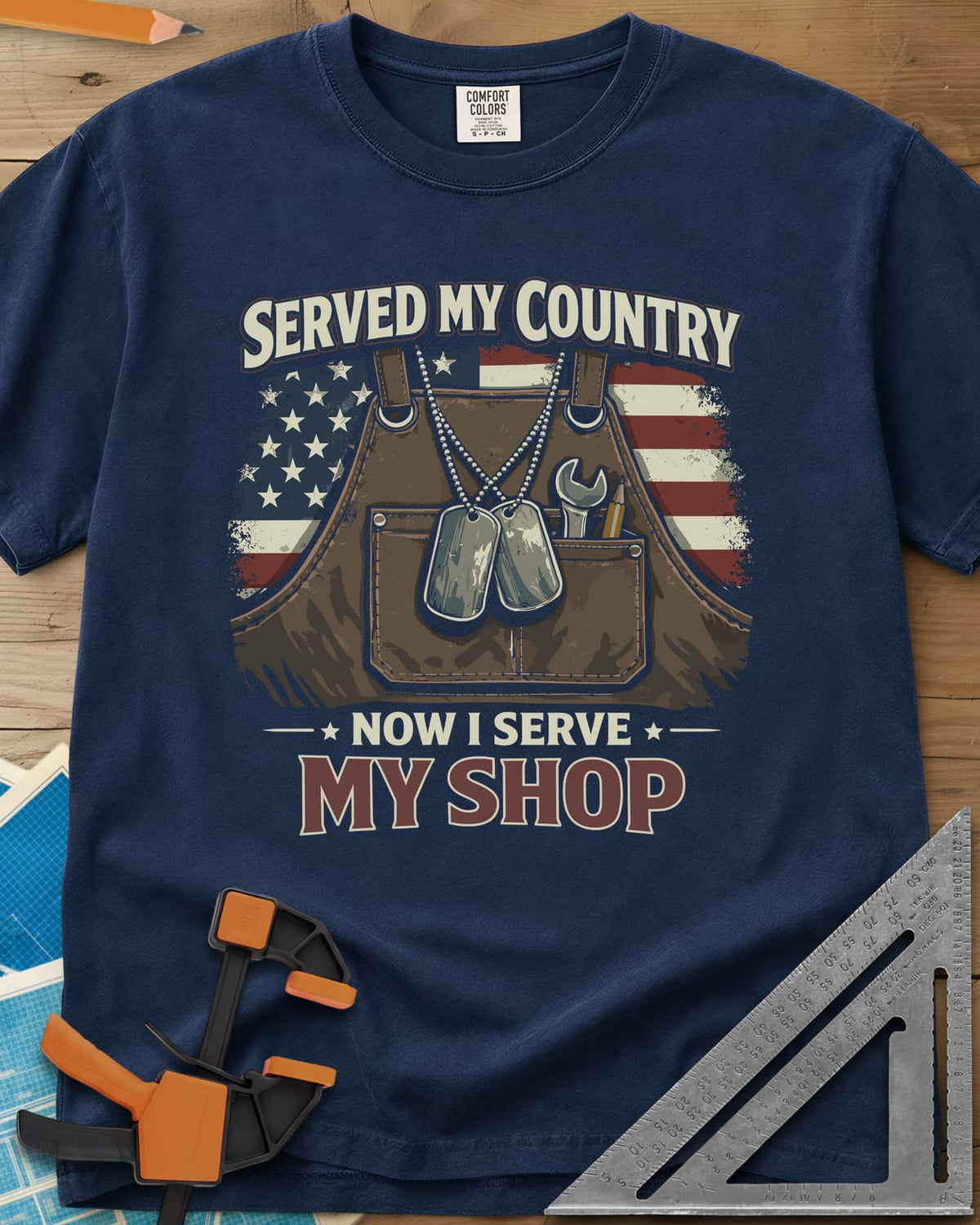 Now I Serve My Shop heavyweight tee in Navy, a shop apron with dog tags, a wrench and a pencil before a flag