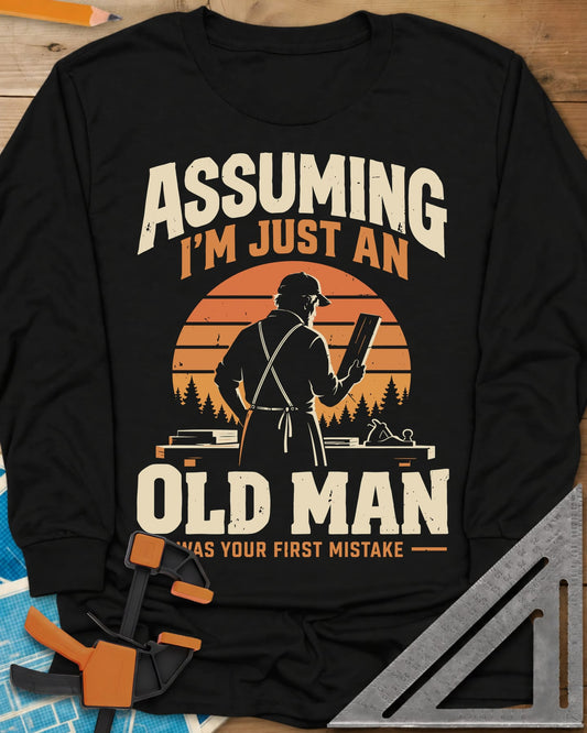 Old Man Mistake long sleeve shirt in Black, a woodworker in an apron holding a board before a striped sunset