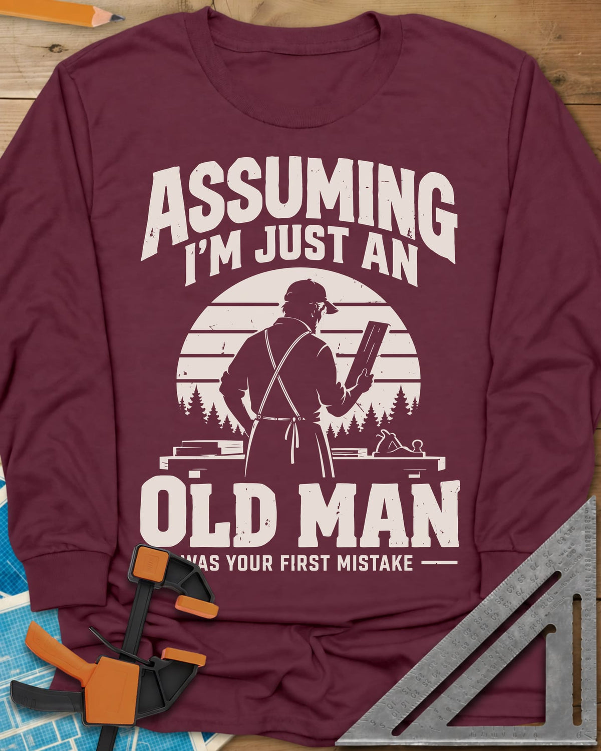 Old Man Mistake long sleeve shirt in Maroon, a woodworker in an apron holding a board before a striped sunset