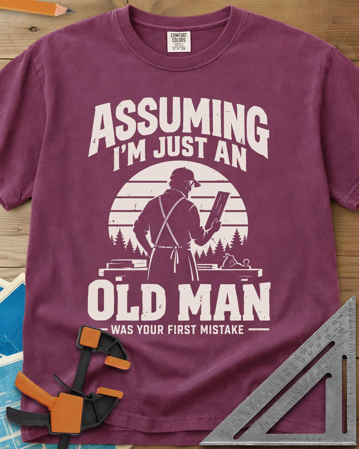 Old Man Mistake heavyweight tee in Berry, a woodworker in an apron holding a board before a striped sunset