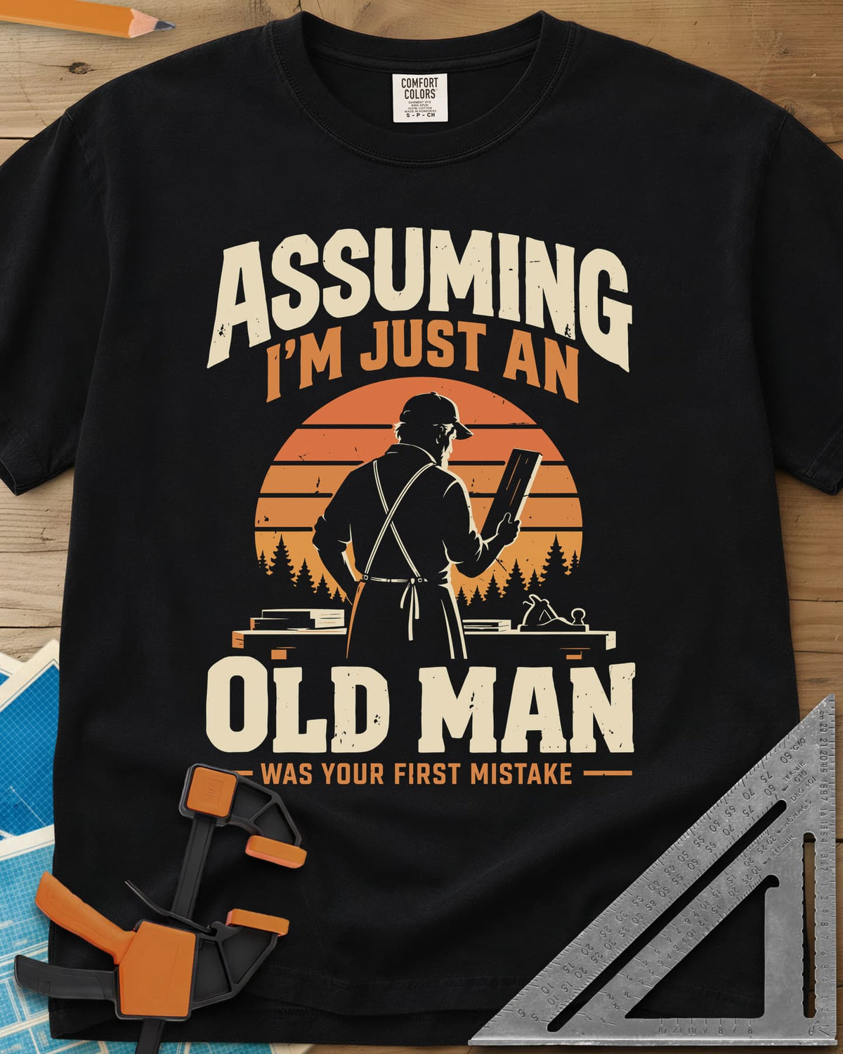 Old Man Mistake heavyweight tee in Black, a woodworker in an apron holding a board before a striped sunset