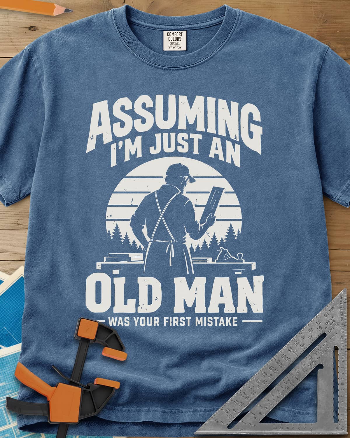 Old Man Mistake heavyweight tee in Blue Jean, a woodworker in an apron holding a board before a striped sunset