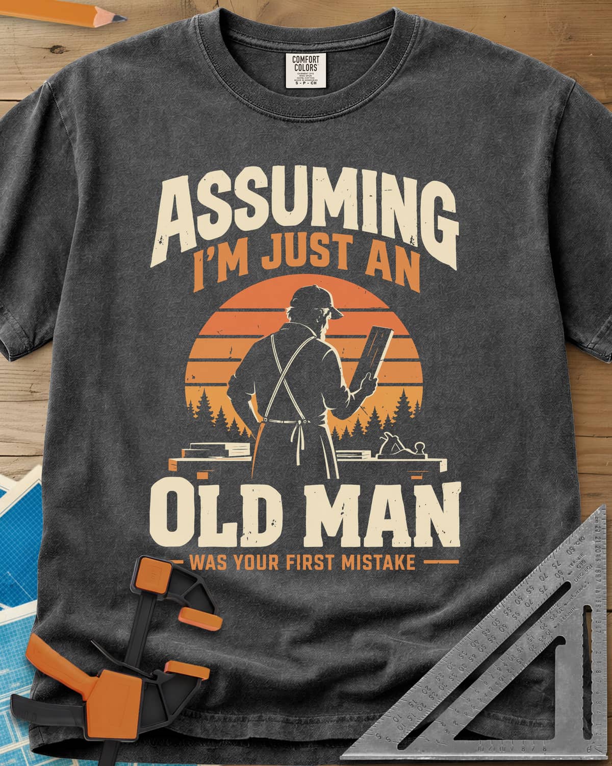 Old Man Mistake heavyweight tee in Pepper, a woodworker in an apron holding a board before a striped sunset