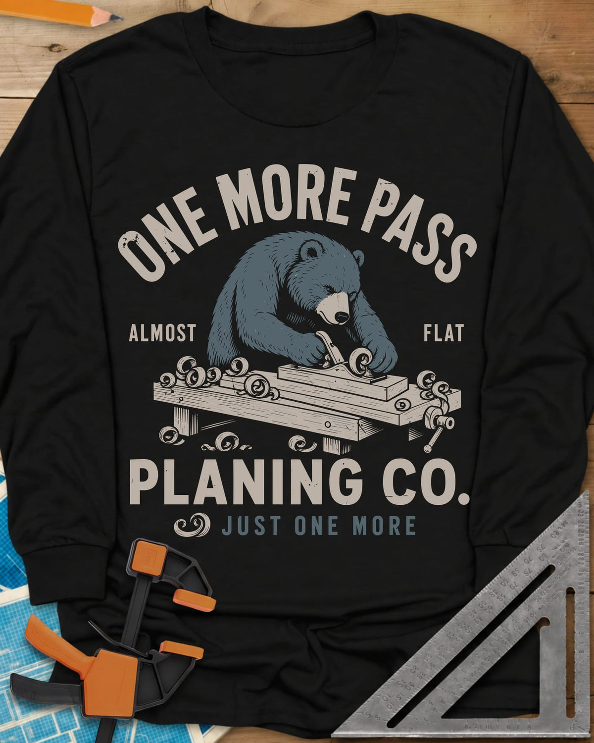 One More Pass long sleeve shirt in Black, a blue bear hand-planing a board at a workbench, shavings curling