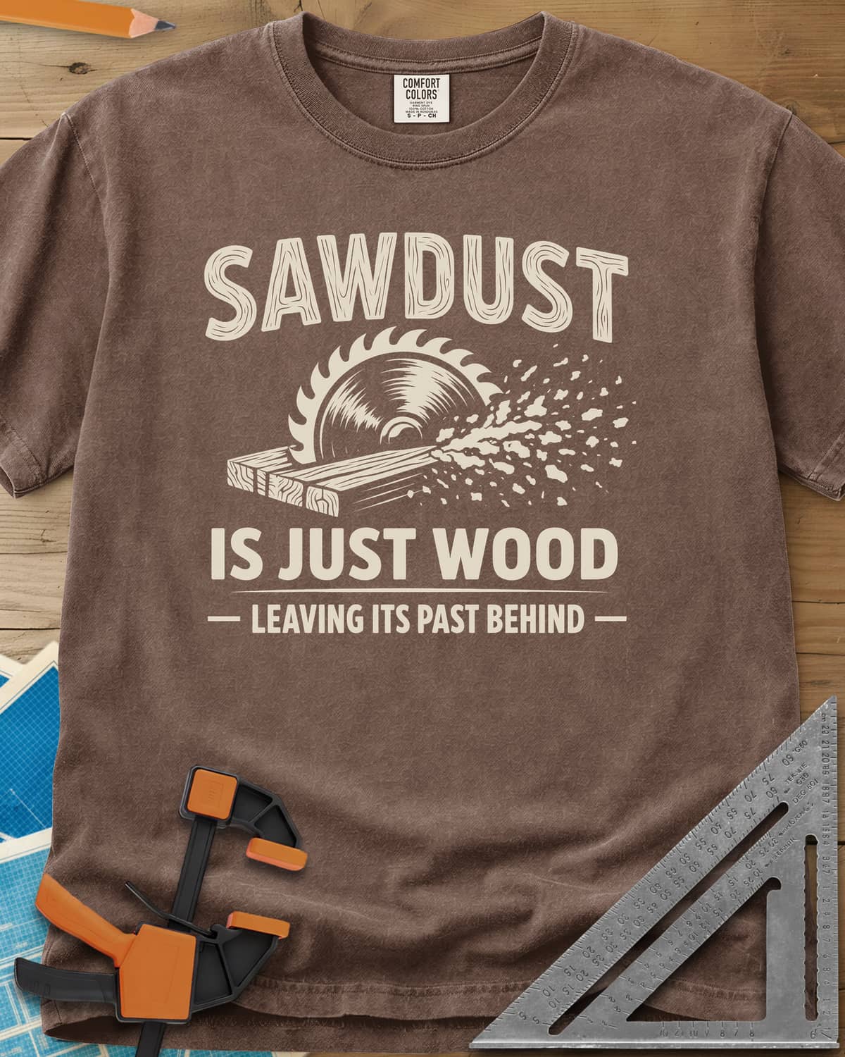 Sawdust Leaving Its Past Behind heavyweight tee in Espresso, wood-grain lettering above a circular saw cutting a board