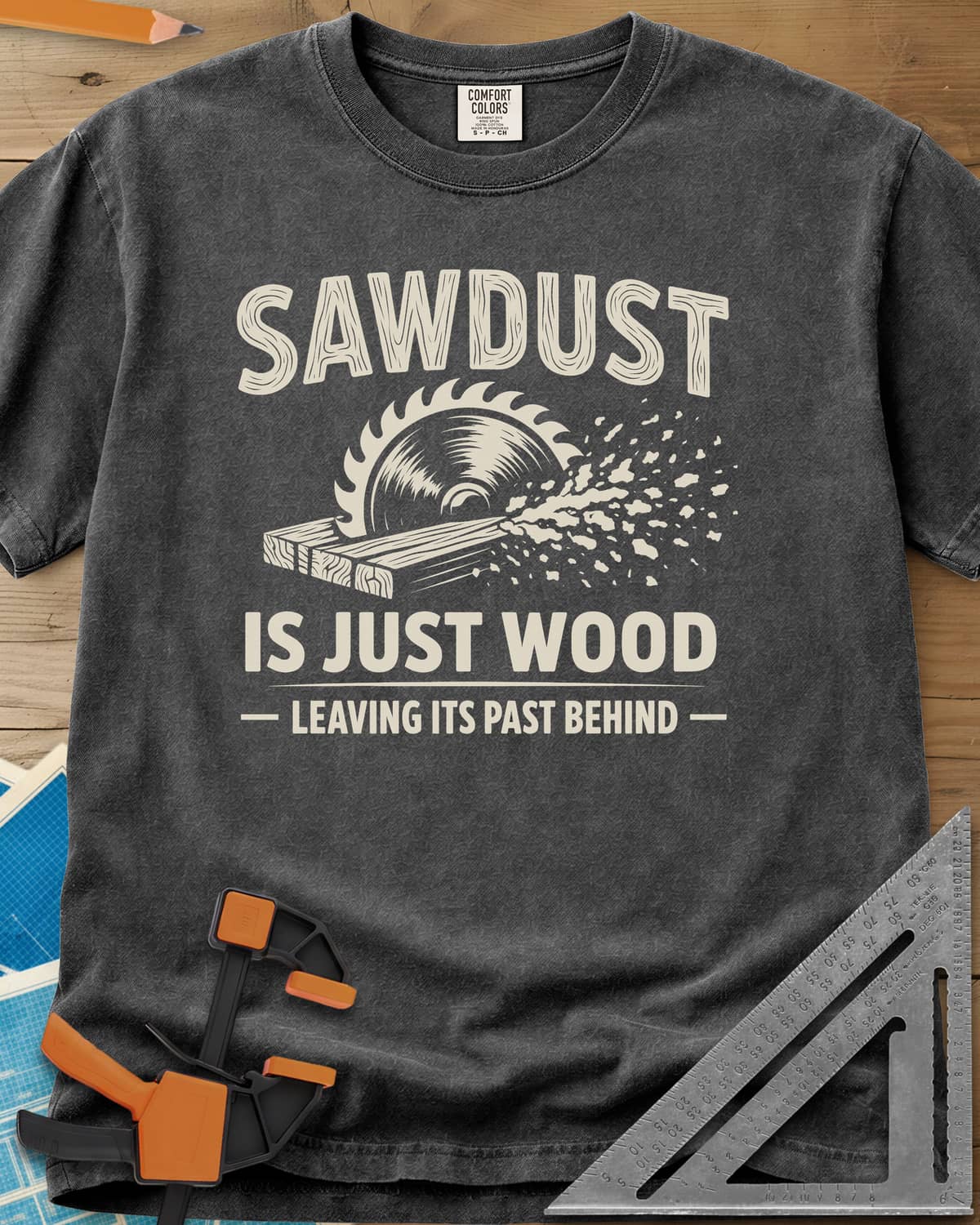 Sawdust Leaving Its Past Behind heavyweight tee in Pepper, wood-grain lettering above a circular saw cutting a board