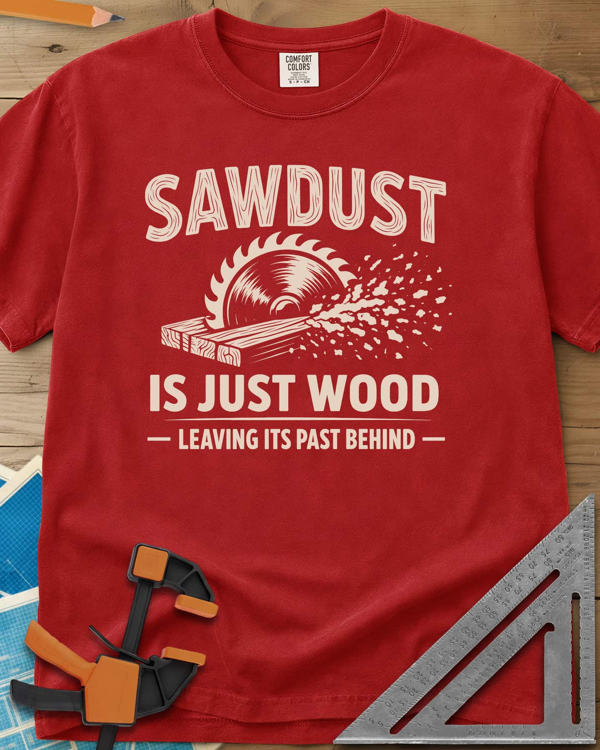 Sawdust Leaving Its Past Behind heavyweight tee in Red, wood-grain lettering above a circular saw cutting a board