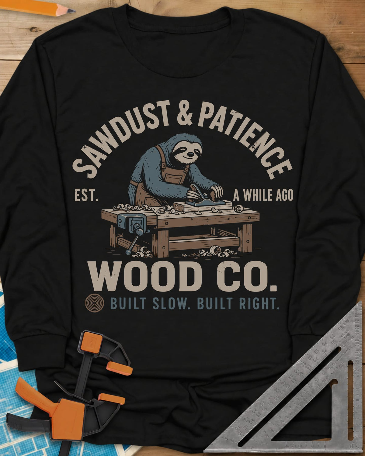 Sawdust & Patience long sleeve shirt in Black, a sloth in an apron planing wood at a bench with a vise