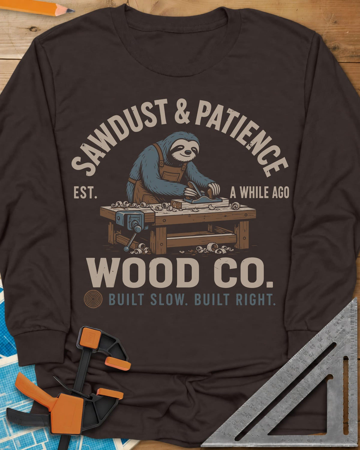 Sawdust & Patience long sleeve shirt in Dark Chocolate, a sloth in an apron planing wood at a bench with a vise