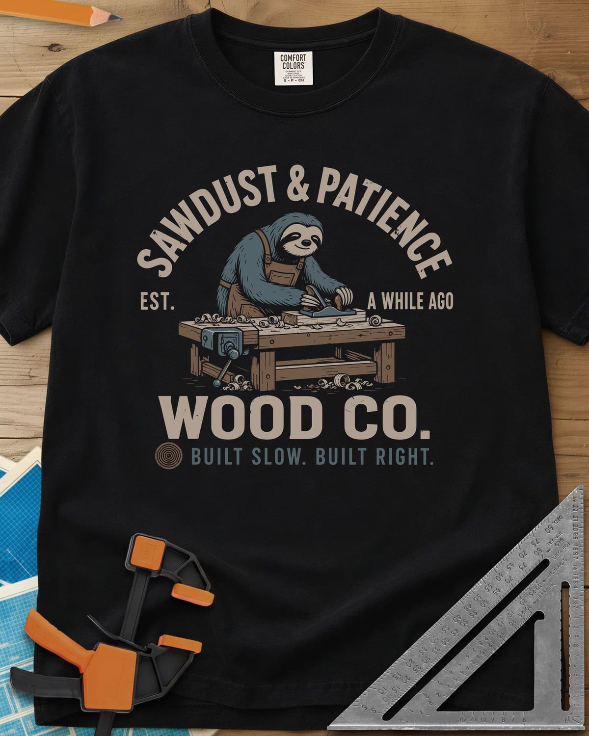 Sawdust & Patience heavyweight tee in Black, a sloth in an apron planing wood at a bench with a vise