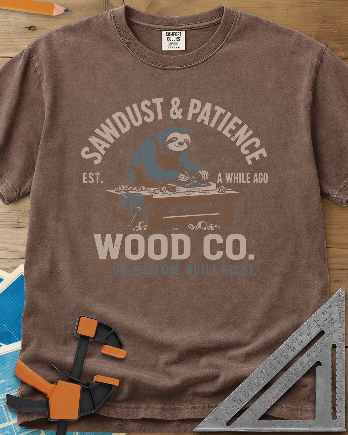 Sawdust & Patience heavyweight tee in Espresso, a sloth in an apron planing wood at a bench with a vise
