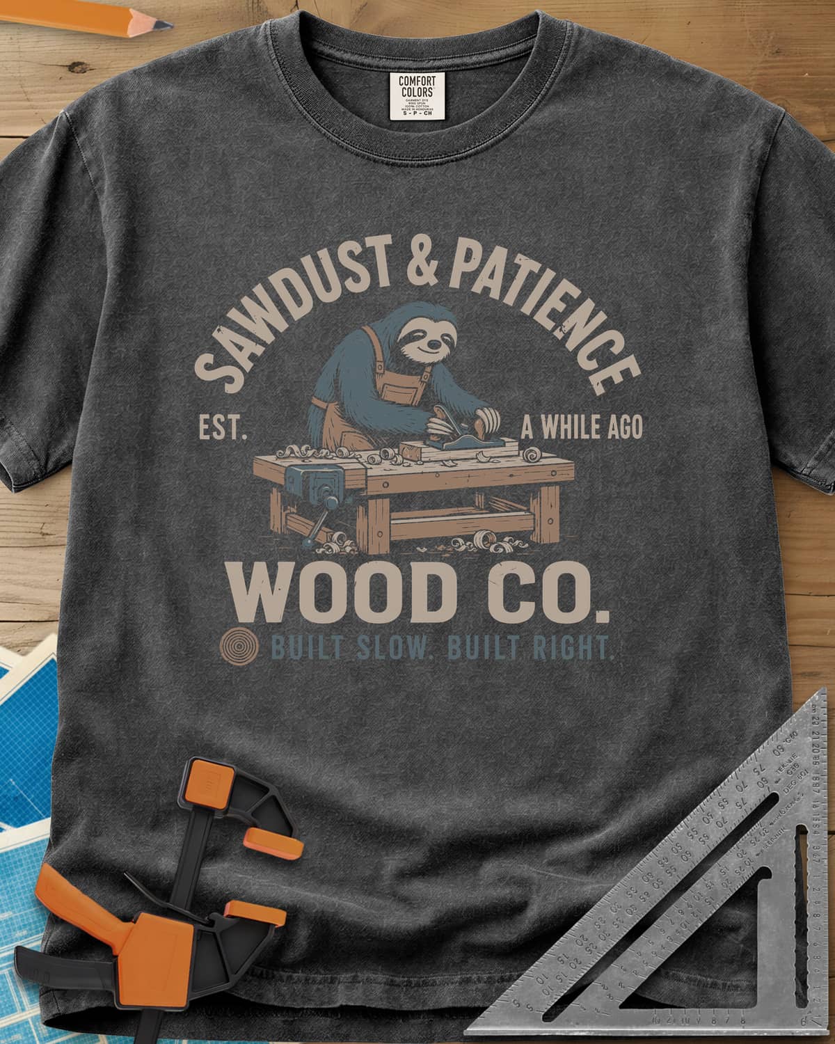 Sawdust & Patience heavyweight tee in Pepper, a sloth in an apron planing wood at a bench with a vise