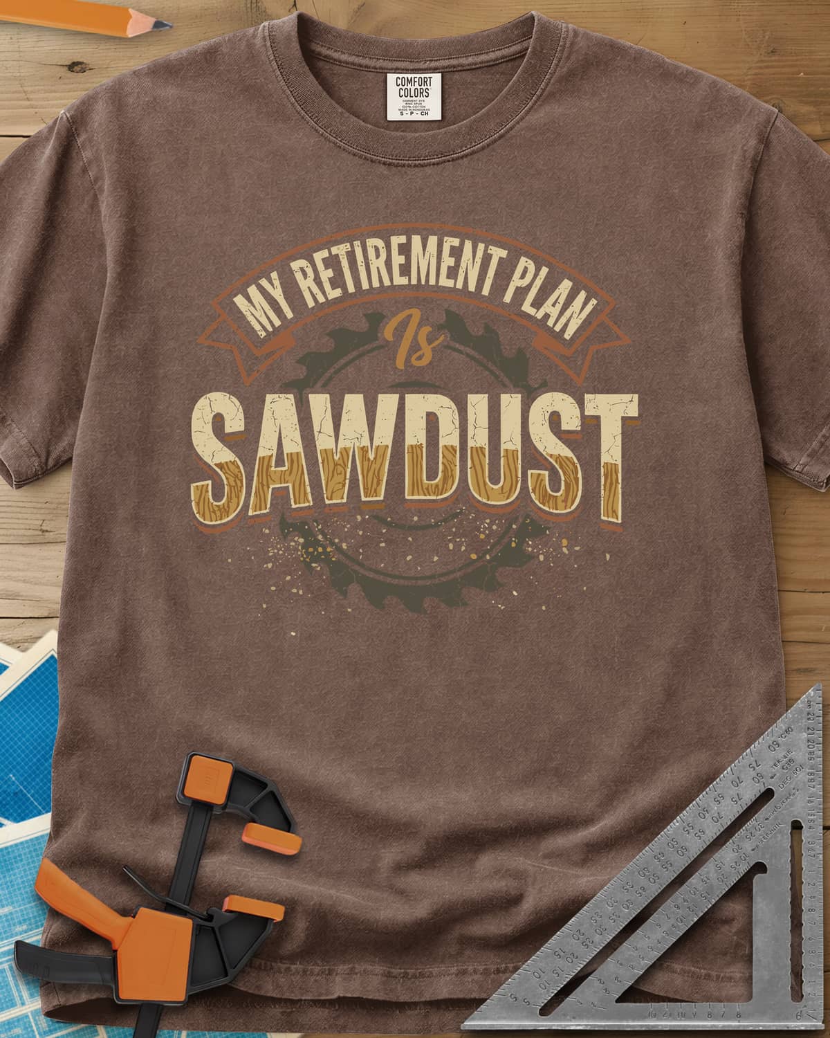 Sawdust Retirement heavyweight tee in Espresso, a ribbon banner over a saw blade, cracked wood-grain letters