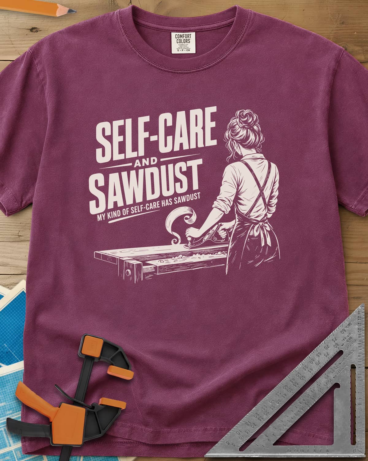 Self-Care & Sawdust heavyweight tee in Berry, a woman at a workbench running a hand plane, shaving curling up