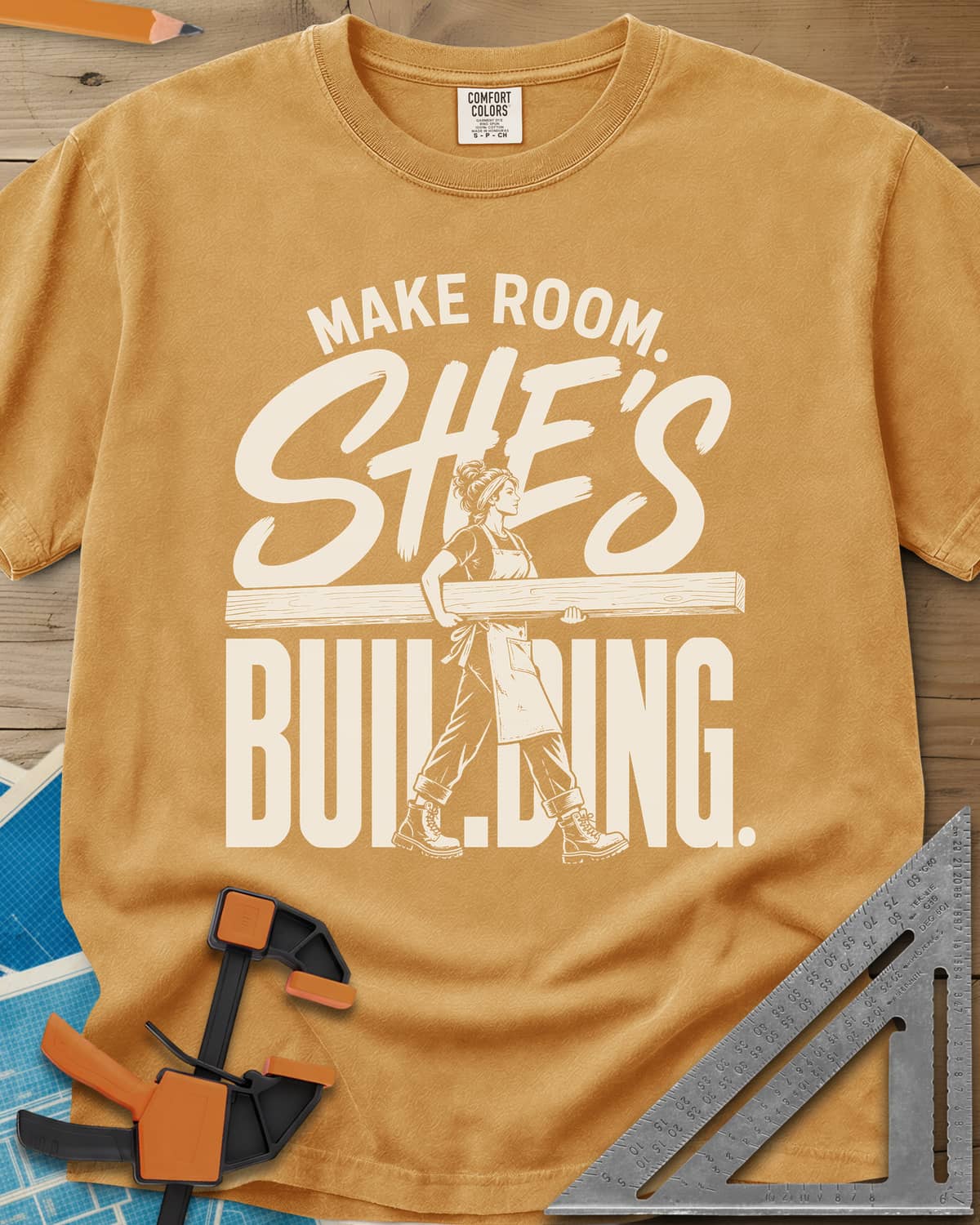 She'S Building heavyweight tee in Mustard, a woman in boots carrying a long beam through the lettering