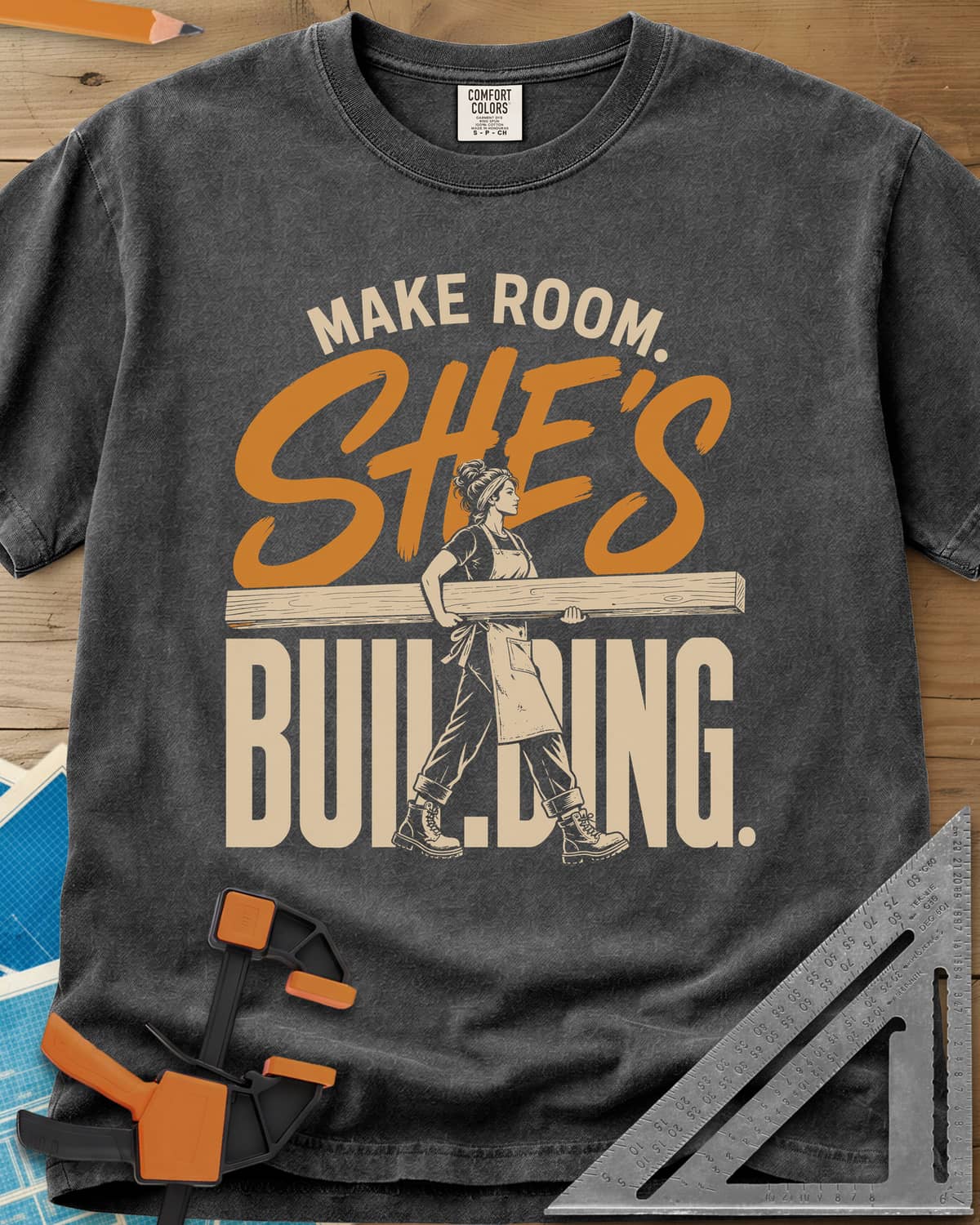 She'S Building heavyweight tee in Pepper, a woman in boots carrying a long beam through the lettering