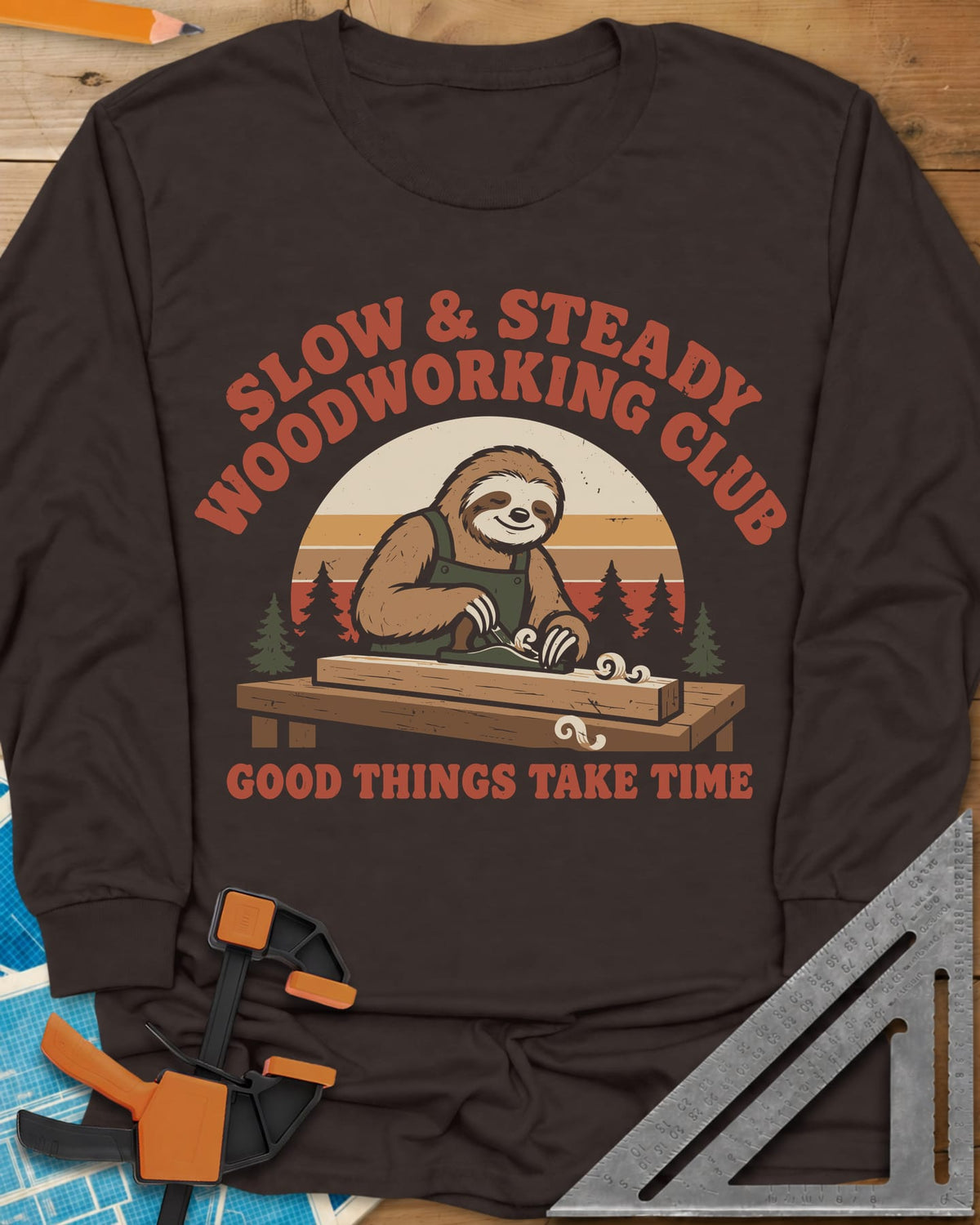 Slow Woodwork long sleeve shirt in Dark Chocolate, a sloth in overalls planing a board, retro sunset and pines