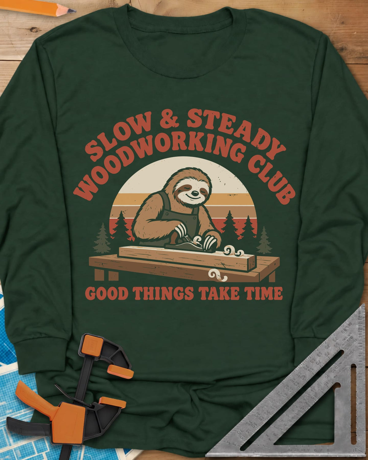 Slow Woodwork long sleeve shirt in Forest Green, a sloth in overalls planing a board, retro sunset and pines