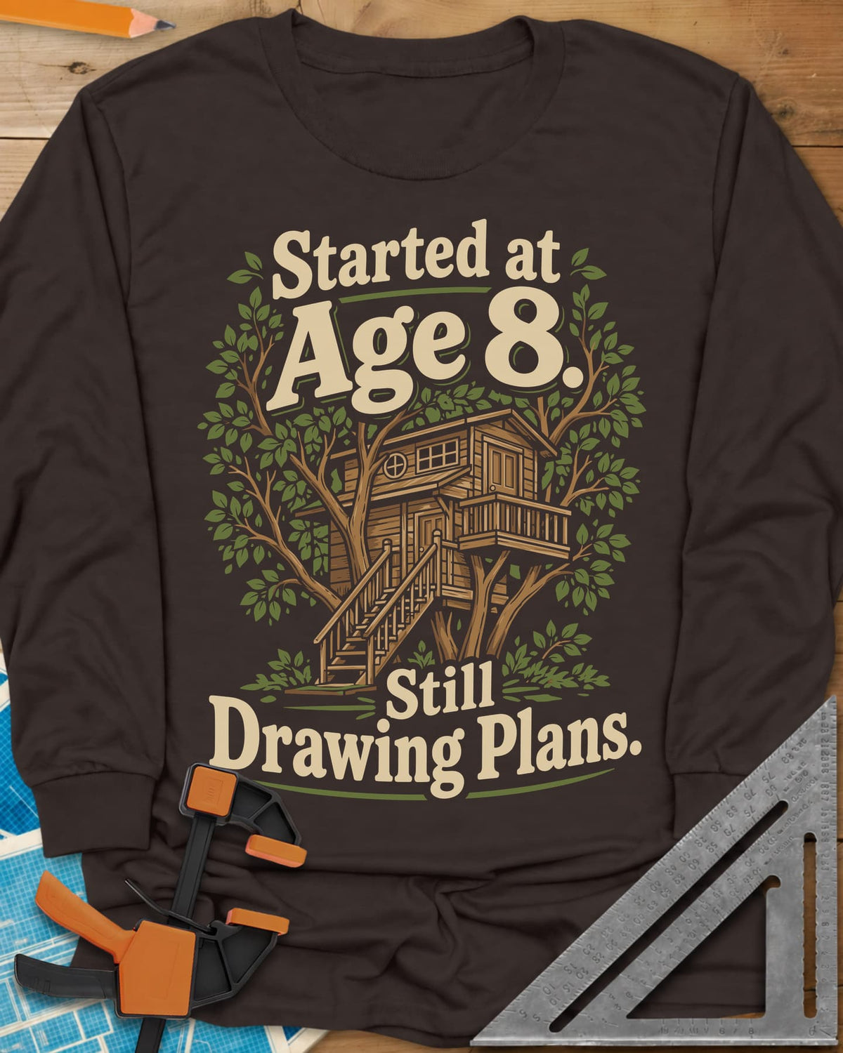 Started At Age 8 long sleeve shirt in Dark Chocolate, a wooden treehouse with a deck and stairs among green leaves