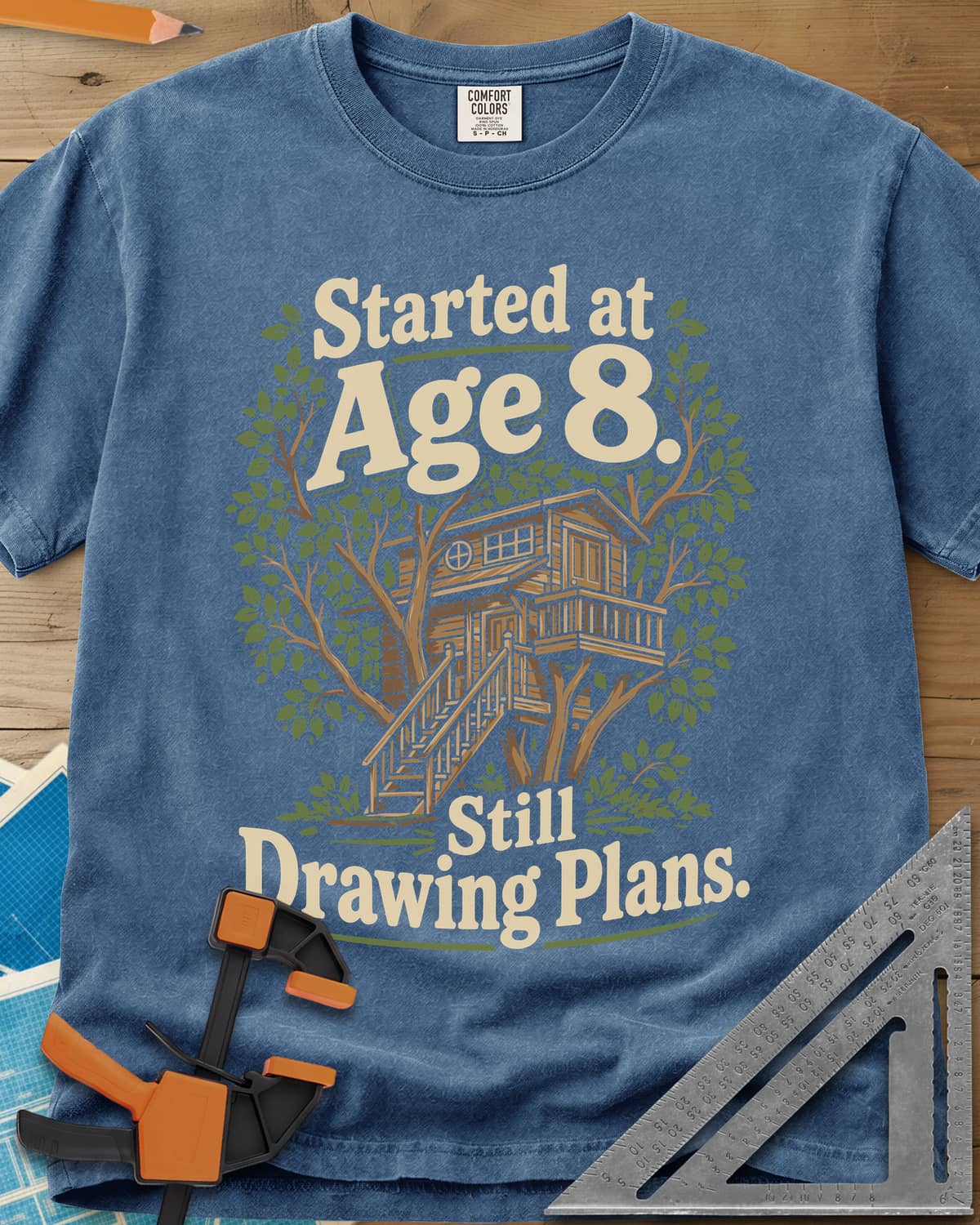 Started At Age 8 heavyweight tee in Blue Jean, a wooden treehouse with a deck and stairs among green leaves