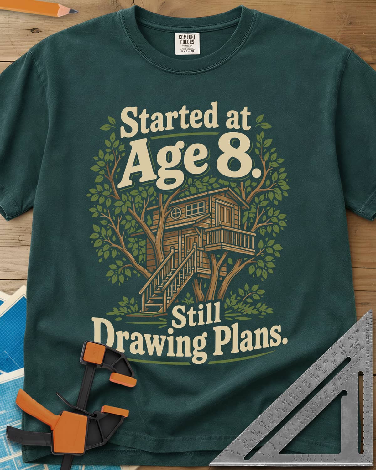 Started At Age 8 heavyweight tee in Blue Spruce, a wooden treehouse with a deck and stairs among green leaves