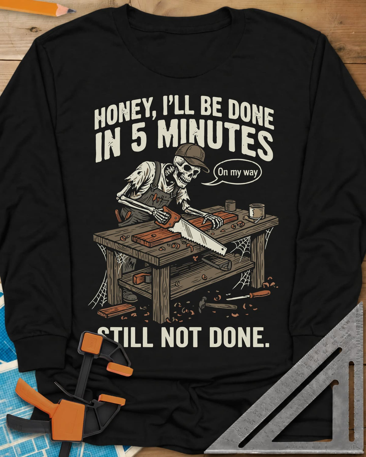 Still Not Done long sleeve shirt in Black, a skeleton in a ball cap sawing a board at a cobwebbed bench