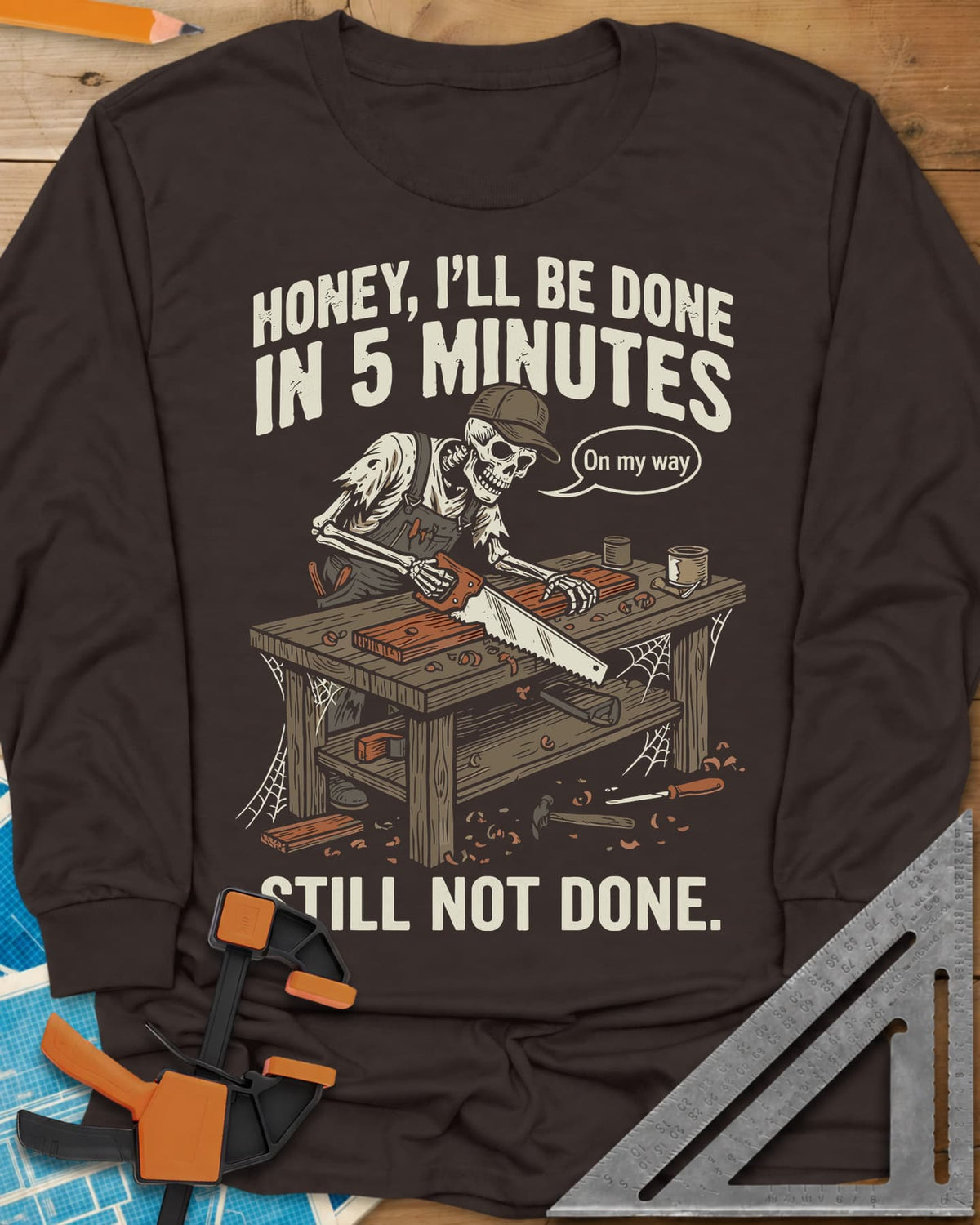 Still Not Done long sleeve shirt in Dark Chocolate, a skeleton in a ball cap sawing a board at a cobwebbed bench
