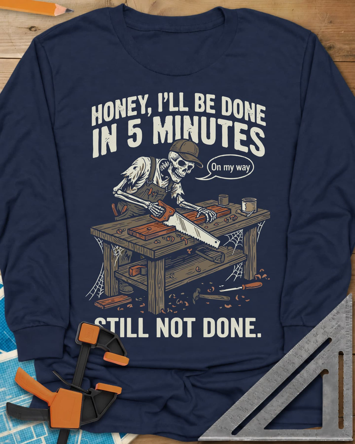 Still Not Done long sleeve shirt in Navy, a skeleton in a ball cap sawing a board at a cobwebbed bench