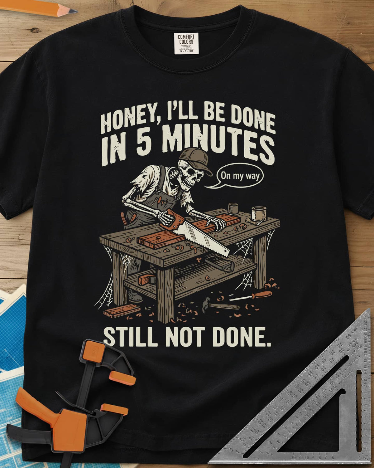 Still Not Done heavyweight tee in Black, a skeleton in a ball cap sawing a board at a cobwebbed bench