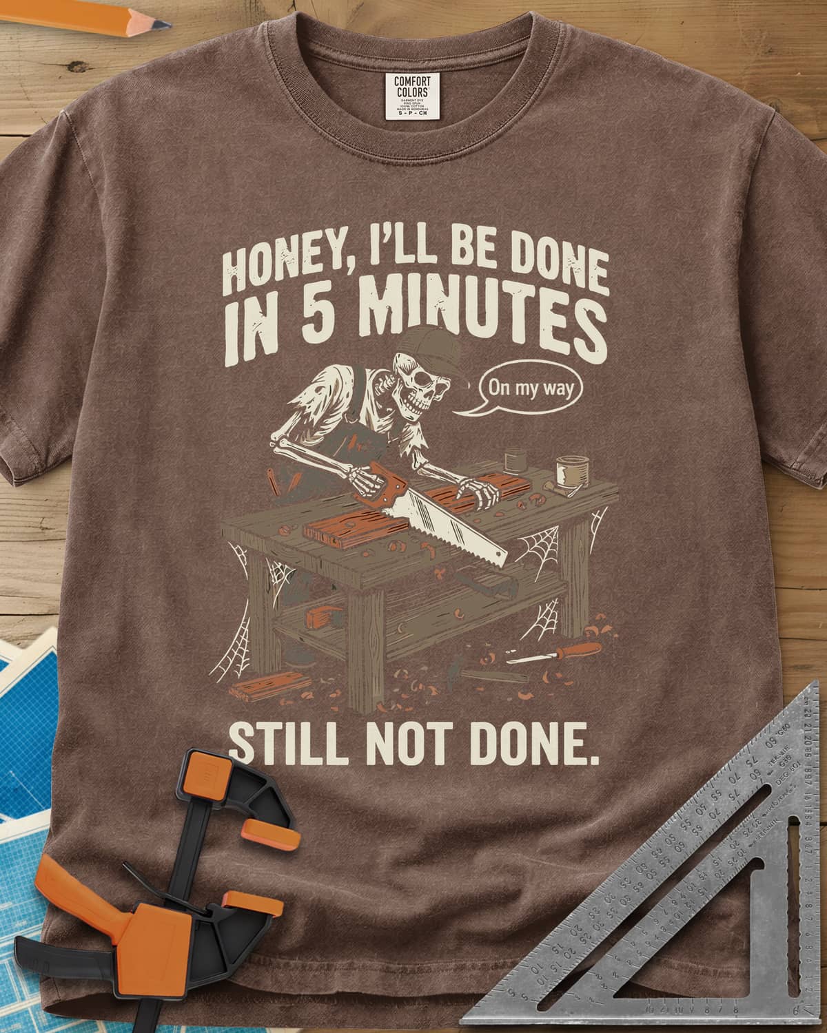 Still Not Done heavyweight tee in Espresso, a skeleton in a ball cap sawing a board at a cobwebbed bench