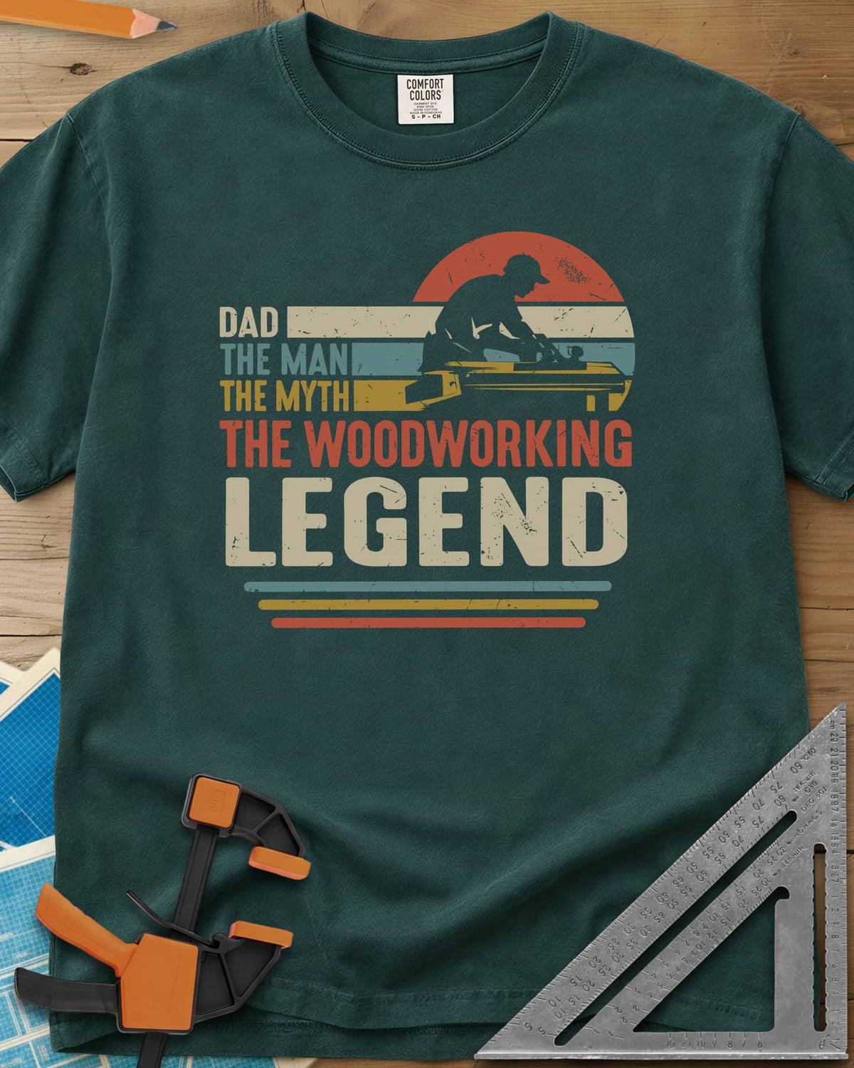 The Woodworking Legend heavyweight tee in Blue Spruce, retro sunset stripes behind a silhouetted woodworker at his bench