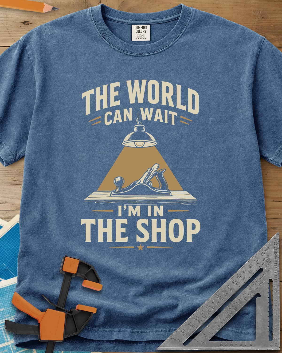 The World Can Wait I'm In The Shop heavyweight tee in Blue Jean, a hand plane under a hanging shop lamp's cone of light
