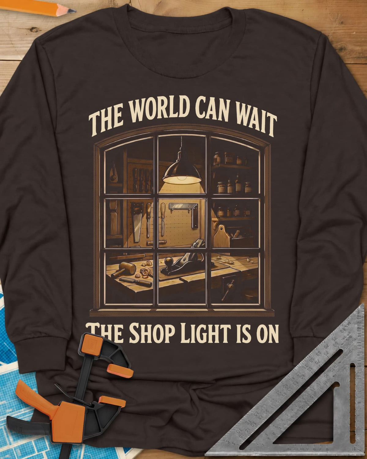 The World Can Wait The Shop Light Is On long sleeve shirt in Dark Chocolate, a lit workshop window: hanging lamp, pegboard