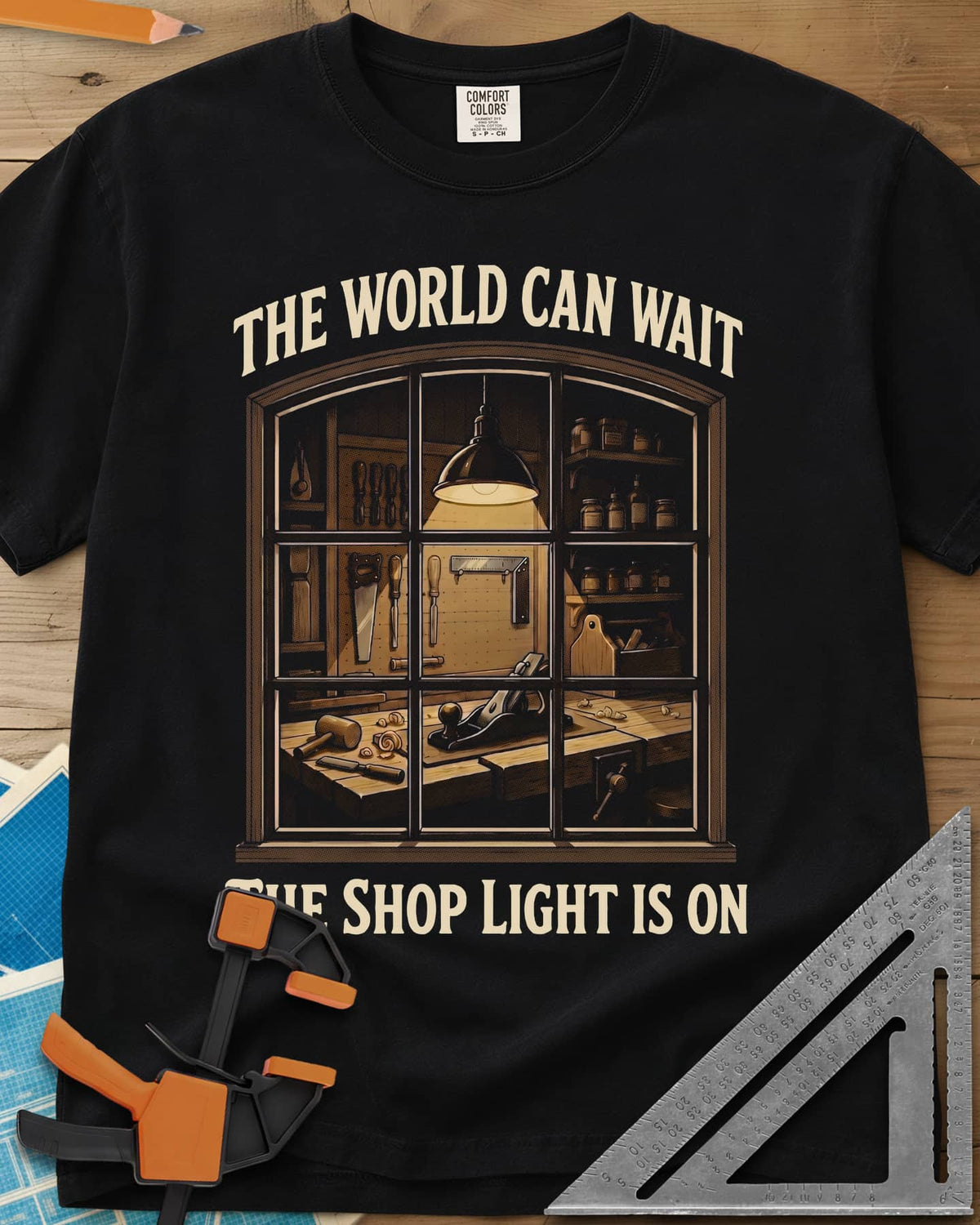 The World Can Wait The Shop Light Is On heavyweight tee in Black, a lit workshop window: hanging lamp, pegboard, hand plane