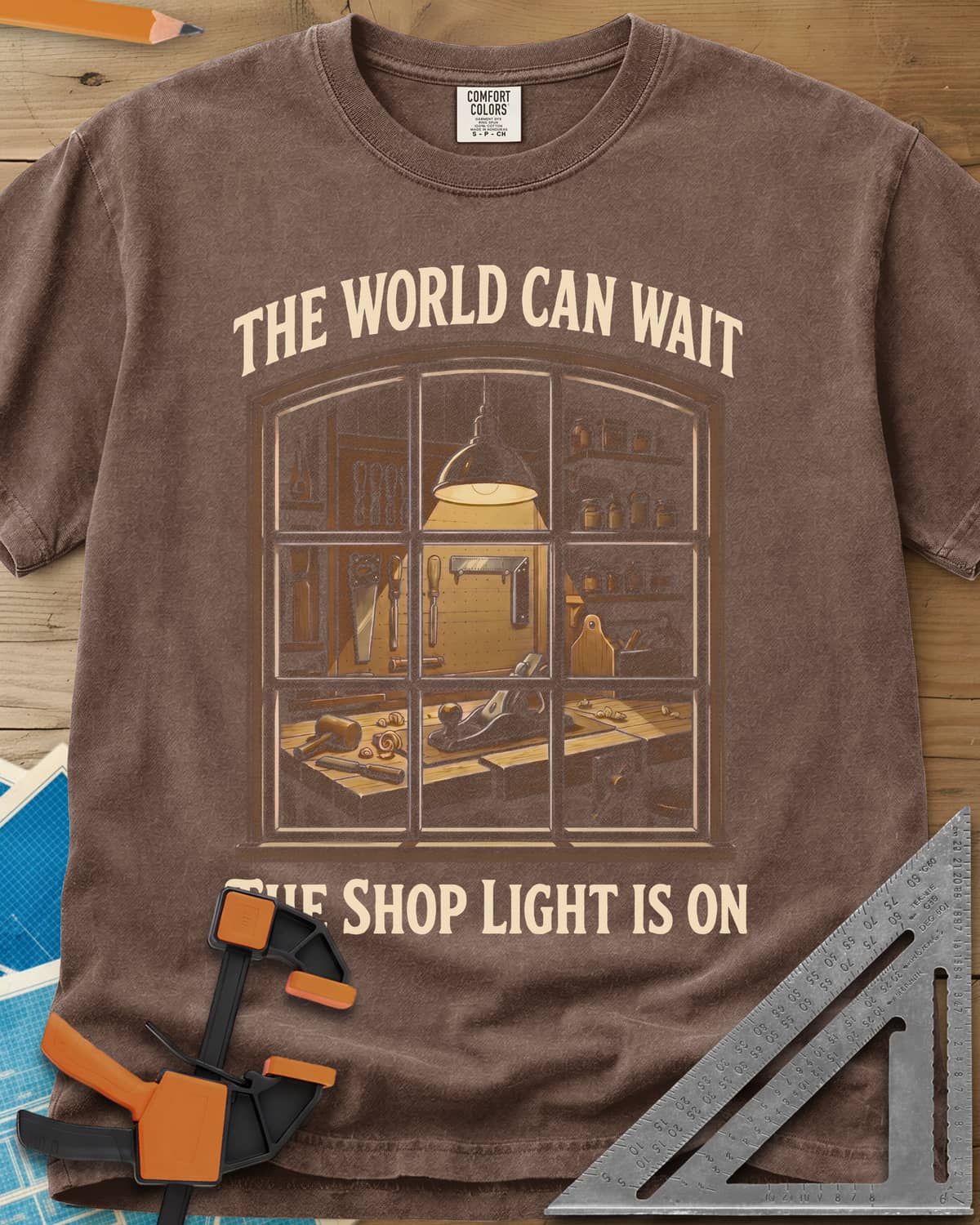 The World Can Wait The Shop Light Is On heavyweight tee in Espresso, a lit workshop window: hanging lamp, pegboard, hand