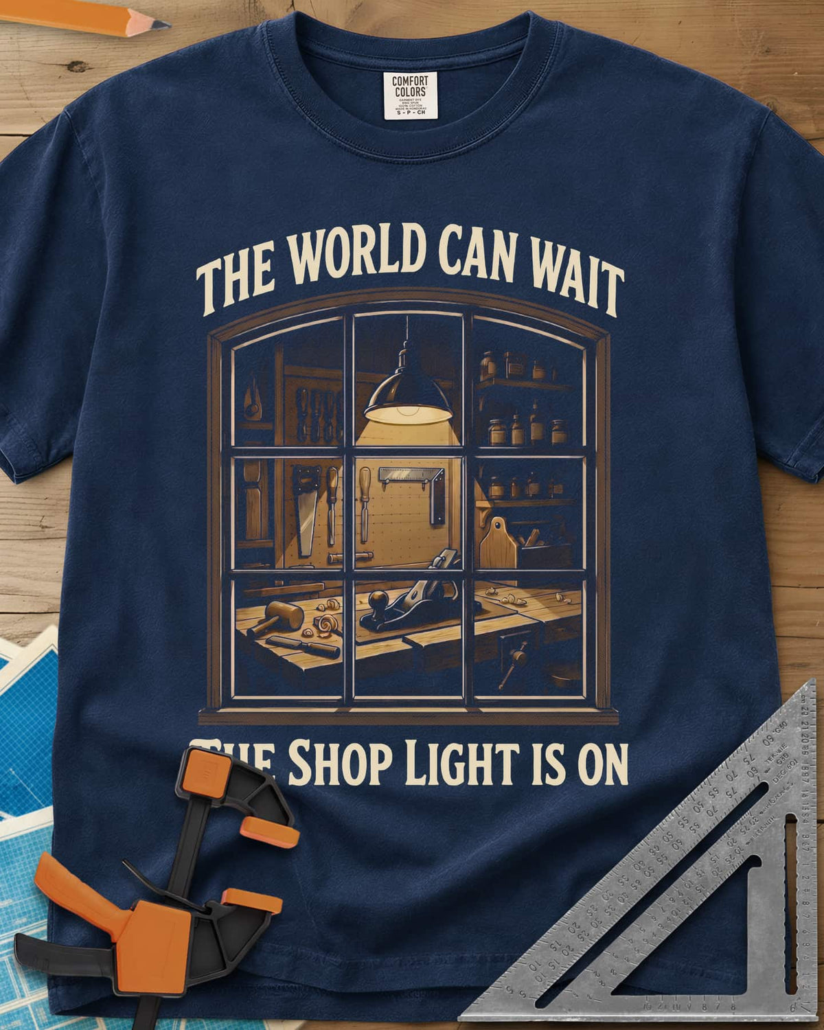 The World Can Wait The Shop Light Is On heavyweight tee in Navy, a lit workshop window: hanging lamp, pegboard, hand plane