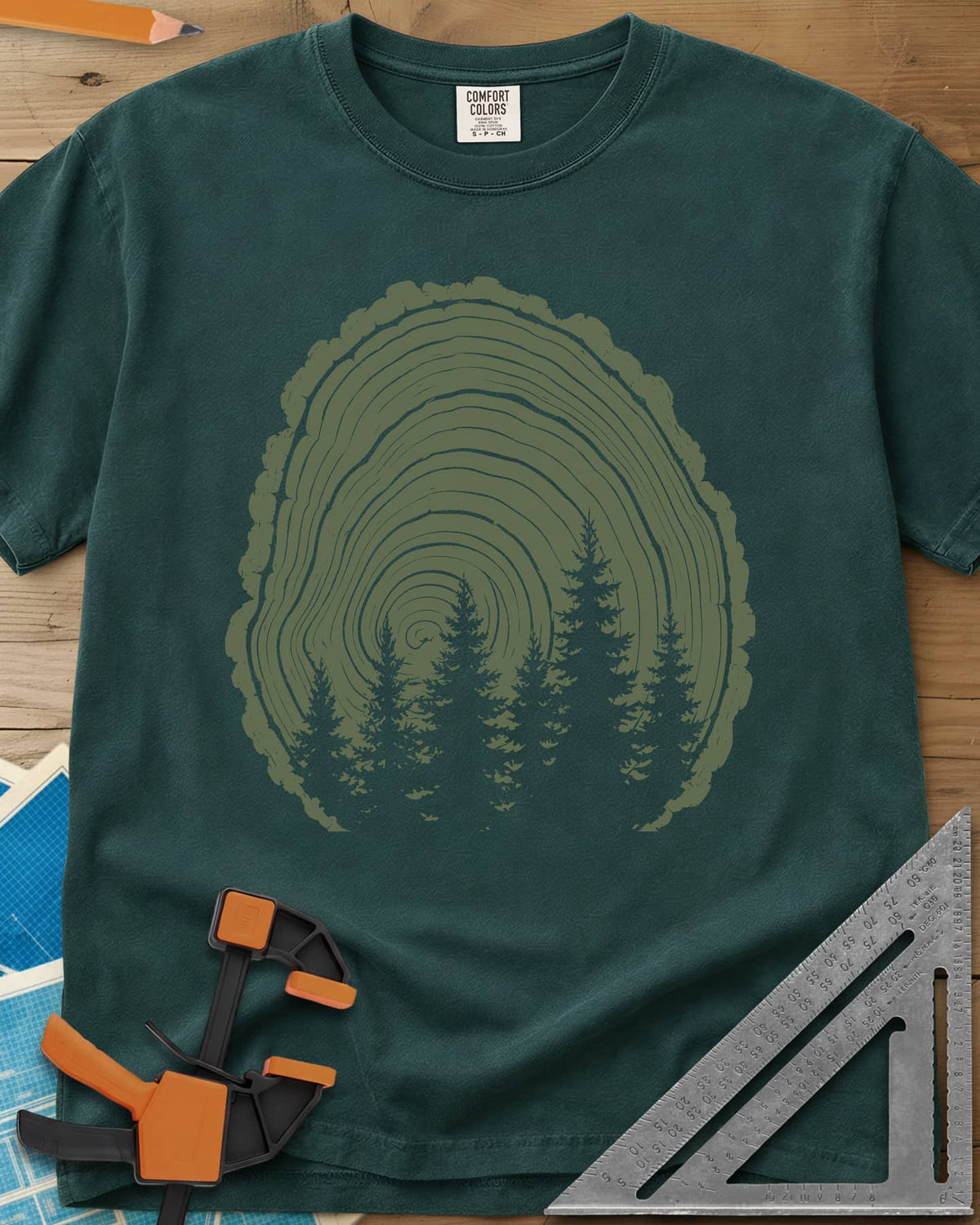 Wood Grain Forest heavyweight tee in Blue Spruce, cut log tree rings with a stand of pine trees inside