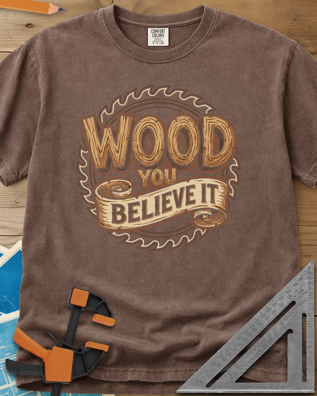 Wood You Believe heavyweight tee in Espresso, wood-grain letters on a scroll banner over a circular saw blade