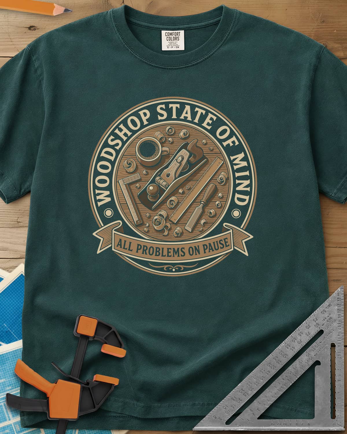 Woodshop State Of Mind heavyweight tee in Blue Spruce, a round badge of a hand plane, coffee, chisel and shavings on a bench
