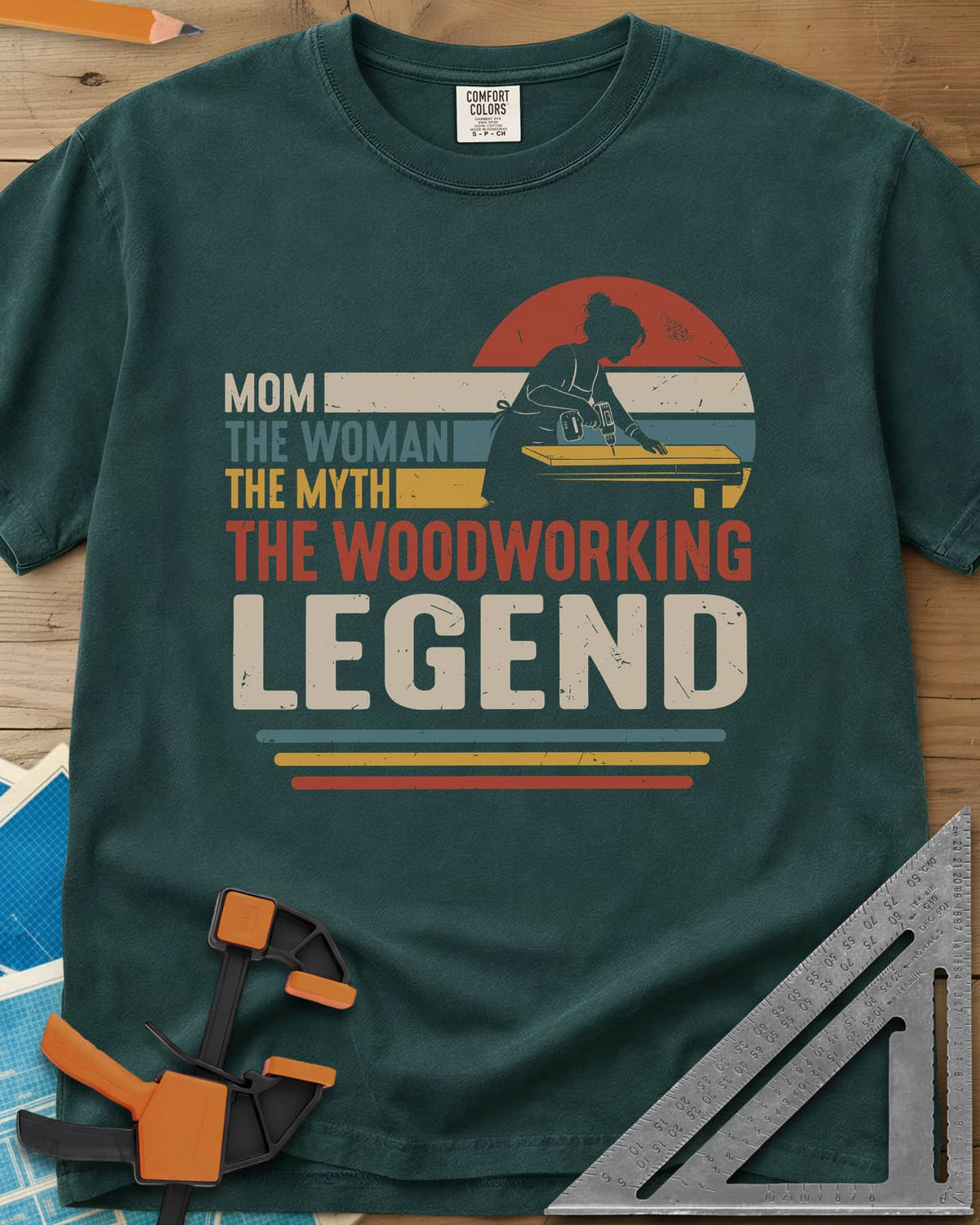 Woodworking Mom Legend heavyweight tee in Blue Spruce, a woman drilling into a board in front of a retro sunset